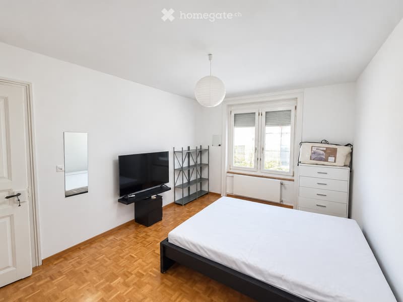 Bright bedroom with wooden flooring, a bed with white bedding, a TV on a stand, a mirror, a metal shelf, and a white dresser with a folded blanket on top.