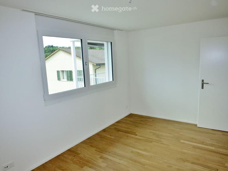 Empty room with white walls, wooden flooring, and a large window offering a view of neighboring houses.