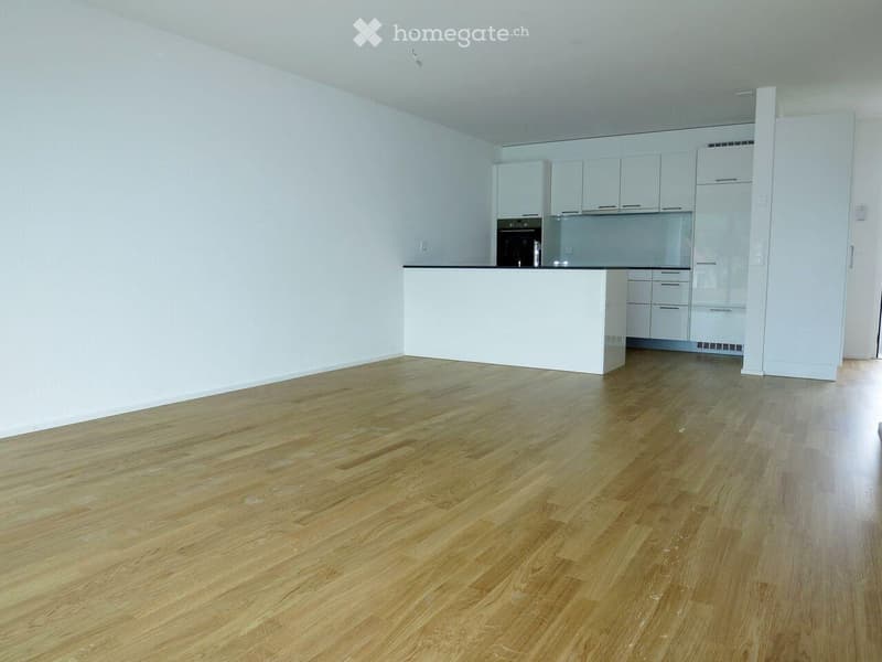 Empty room with light wooden flooring and white walls, featuring a white kitchen area with cabinets and a countertop in the background.