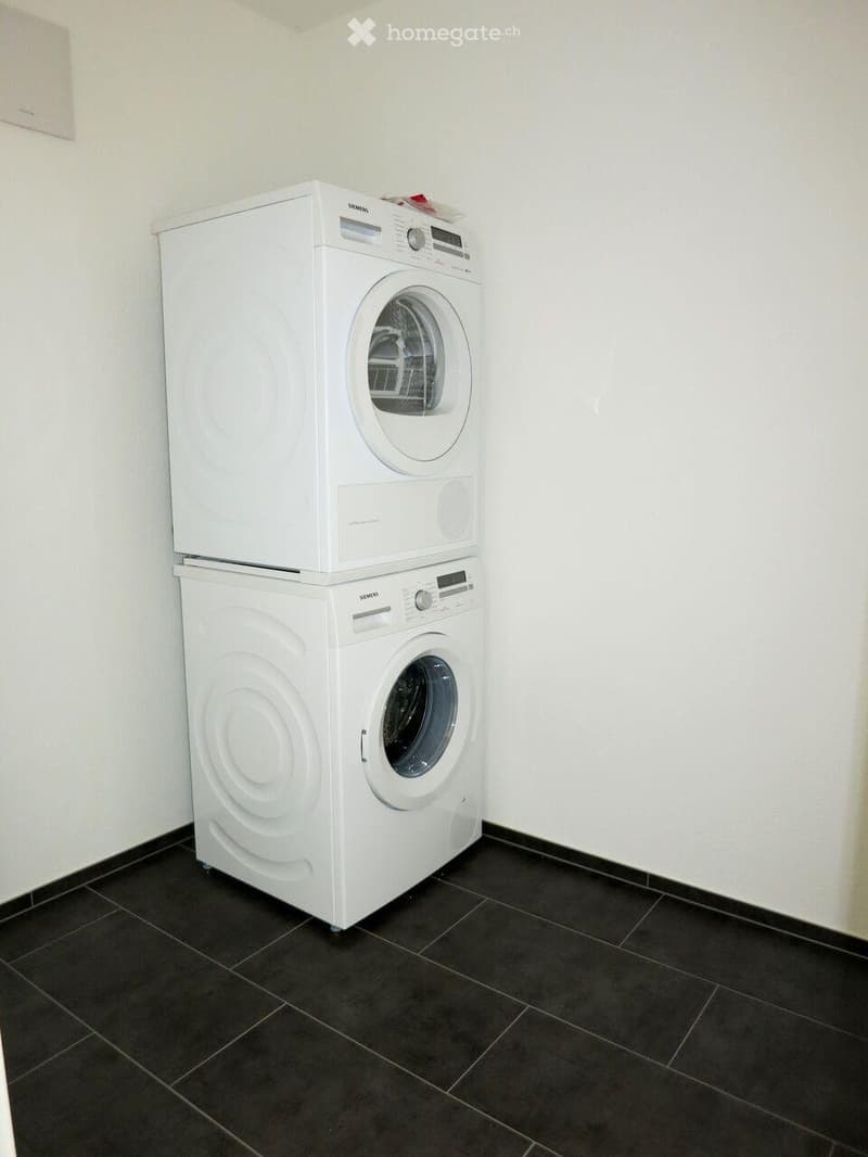 A small room with a black tiled floor and white walls, featuring a stacked washing machine and dryer unit.