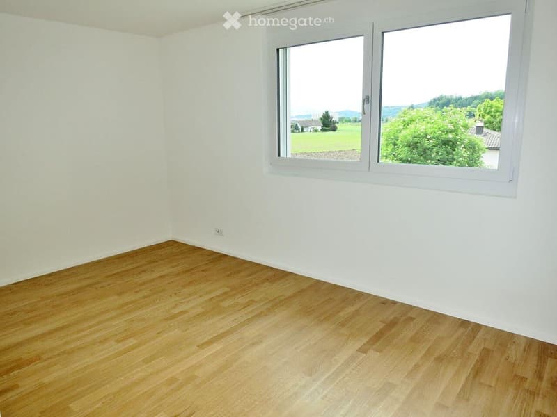 Empty room with light wooden flooring and white walls, featuring large windows with a scenic view of greenery and distant hills.