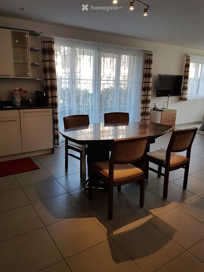 Bright dining area with a round wooden table and four chairs, adjacent to a modern kitchen with white cabinets and a countertop. Large windows with curtains offer a view of the outside.
