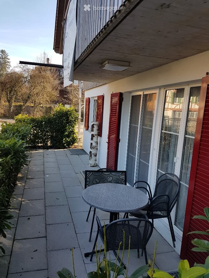 Balcony with a round table and two black metal chairs, surrounded by greenery and a building with red shutters.