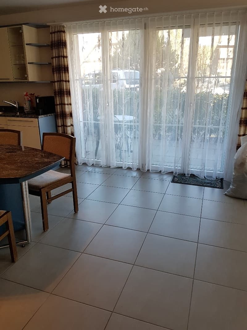 Bright dining area with tiled floor, large window with sheer curtains, round wooden table with chairs, small kitchen area with cabinets and sink.