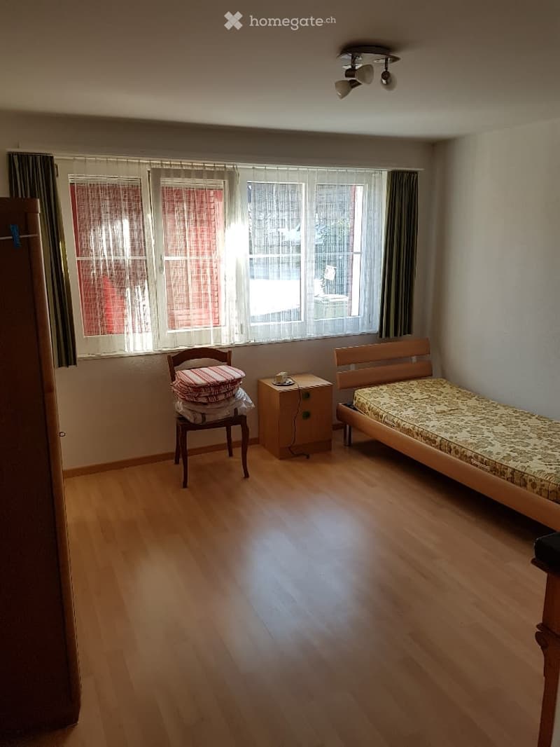 A small, furnished bedroom with wooden flooring, a single bed, a nightstand, a chair with folded blankets, and large windows with curtains.
