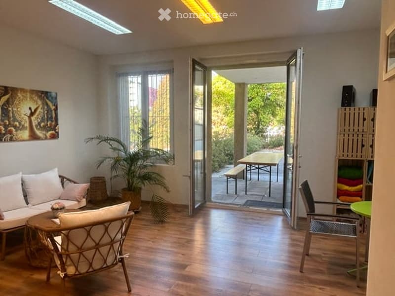 Bright living room with wooden flooring, large windows, and an open sliding door leading to a balcony area with outdoor furniture.