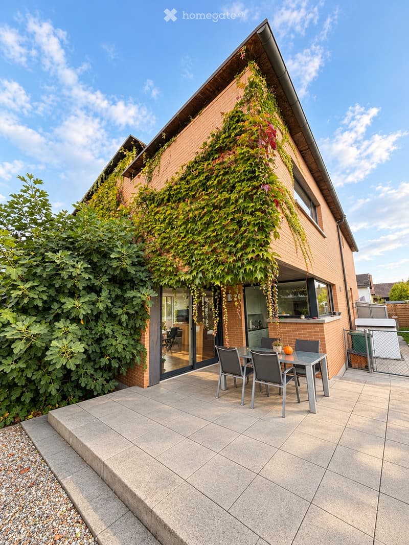A modern patio area with a tiled floor, a small table with four chairs, and a view of the house's living space through large glass doors. The house features a green wall covered in climbing plants.