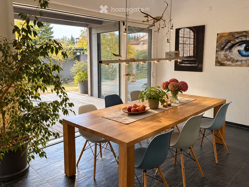 Modern dining room with a wooden table, six chairs, a bowl of fruit, a vase with flowers, and a potted plant. Large windows provide natural light and a view of the garden.