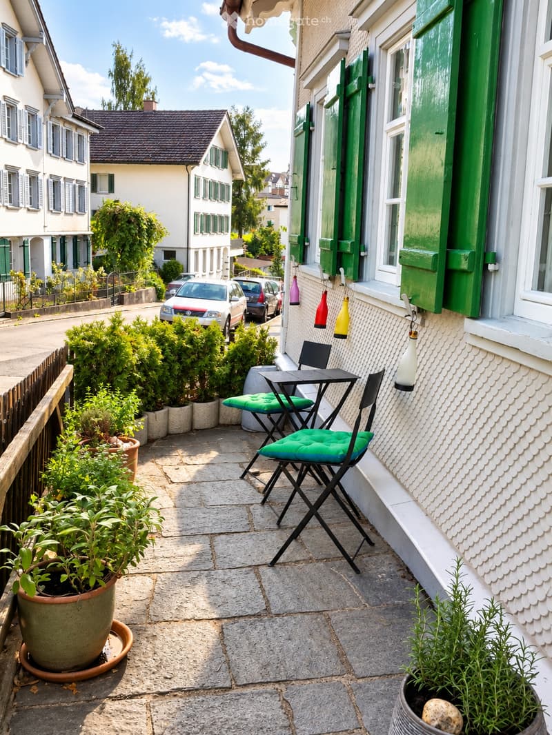 A small balcony with a stone floor, a black folding table, green cushioned chairs, and several potted plants.