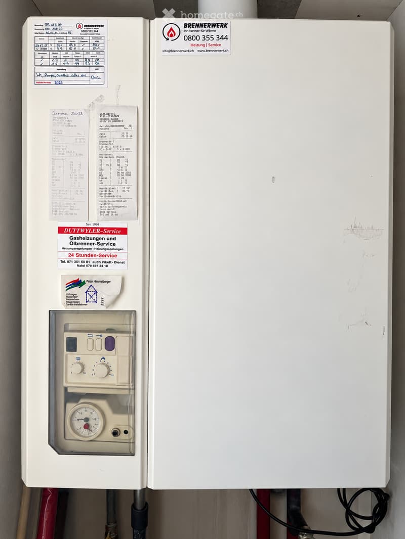 Gas boiler with control panel and gauge, mounted on wall with service stickers and technical data sheets.