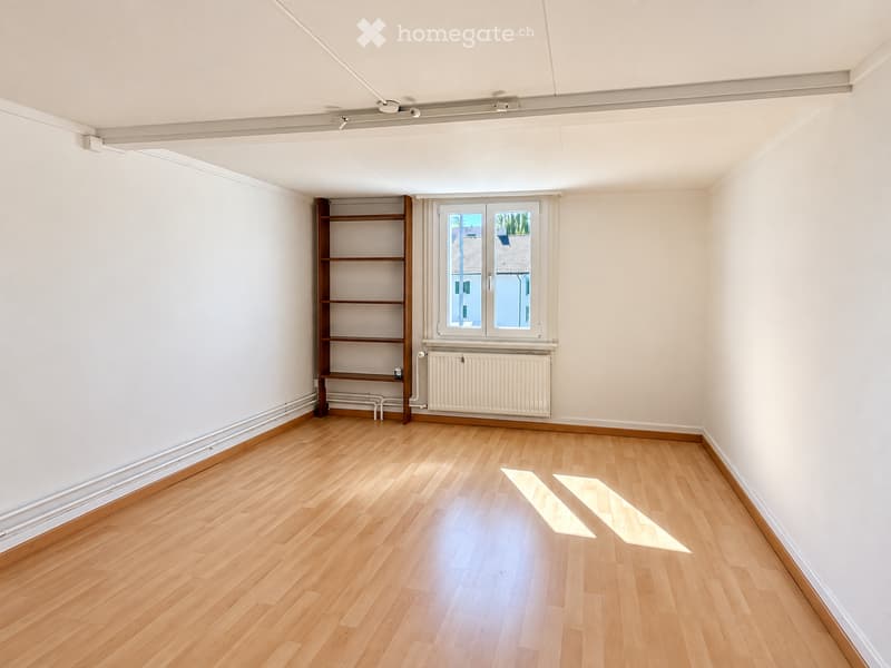 Empty room with light wooden flooring, white walls, a built-in wooden shelf unit, a window with a radiator below, and a ceiling light fixture.