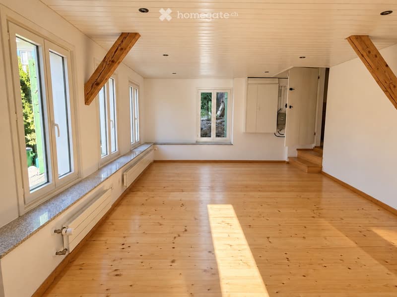 Empty room with wooden flooring, white walls, large windows, and a staircase leading upwards.