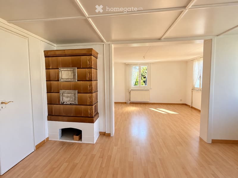 Empty room with light wooden flooring, white walls, and a decorative fireplace on the left side. The room has large windows with white curtains, allowing natural light to fill the space.