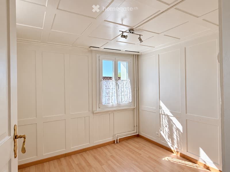Empty room with light wooden flooring, white paneled walls, and a window with white curtains. The ceiling features decorative molding and recessed lighting.