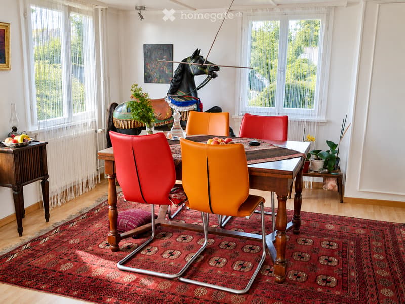 Brightly lit dining area with a wooden table, colorful chairs, a patterned rug, and decorative items including a toy horse, fruit bowl, and paintings.