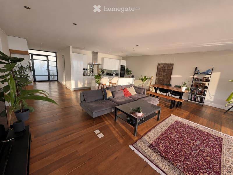 Open-plan living and dining area with wooden flooring, grey sectional sofa, coffee table, dining table with chairs, bookshelf, and potted plants.