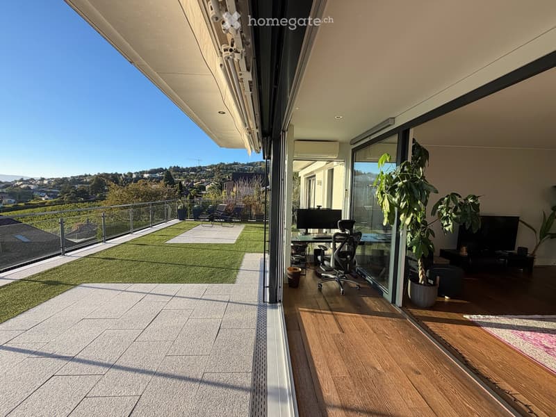 Spacious balcony with wooden flooring, large glass sliding doors, modern furniture including a desk and office chair, potted plants, and a view of a landscaped area with artificial grass and outdoor seating.