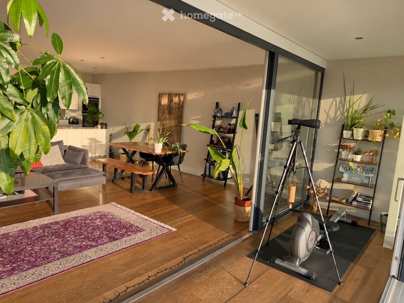 Bright, open living space with wooden flooring, large windows, and modern furniture including a sofa, coffee table, dining table, and exercise bike.