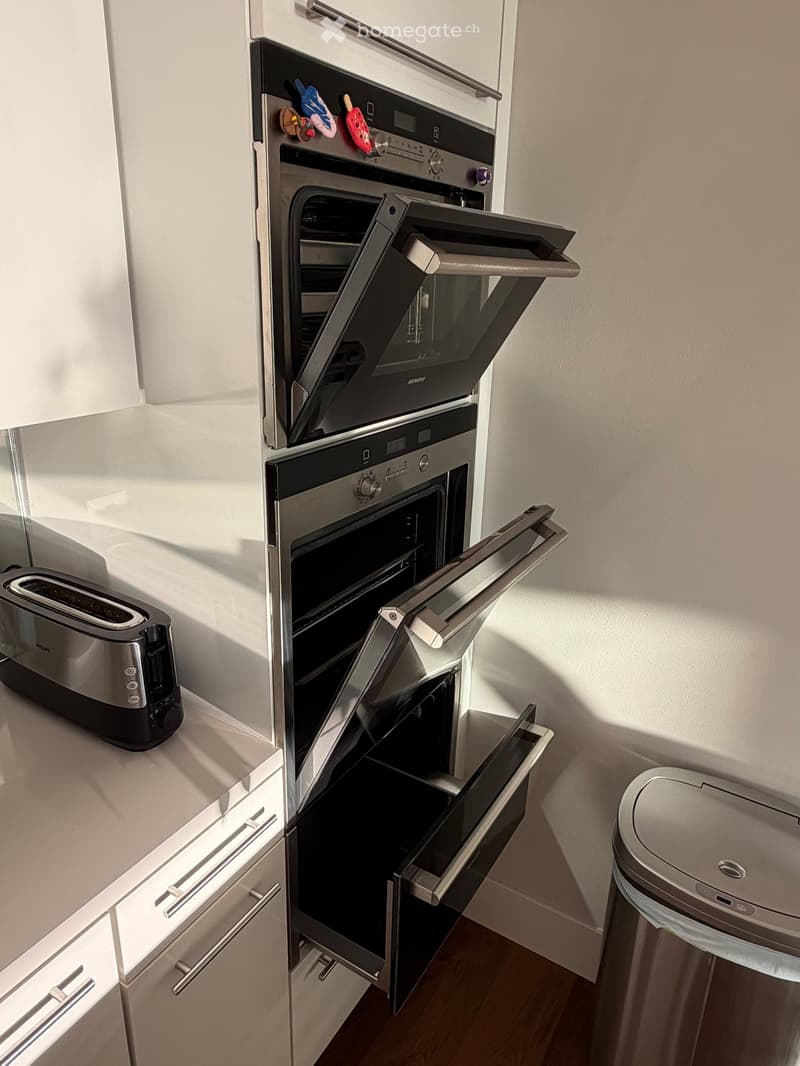 Modern kitchen with stainless steel oven, toaster, and trash bin. The oven doors are open, revealing the interior.