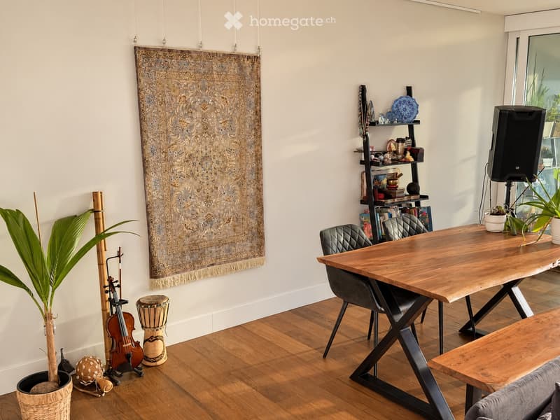 A modern, well-furnished room with a large wooden table, chairs, a shelf with books and decorative items, a speaker, and a large tapestry on the wall. The room also features a potted plant, a drum, and a guitar in the corner.