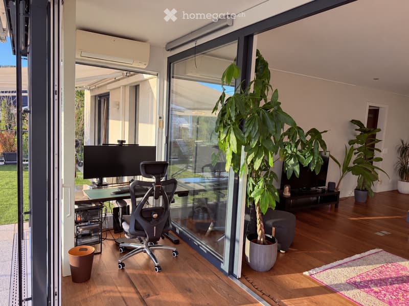 A modern, well-lit room with wooden flooring, large glass windows, and a comfortable office setup including a desk, chair, and computer screen. There are potted plants, a small table, and a trash bin visible.