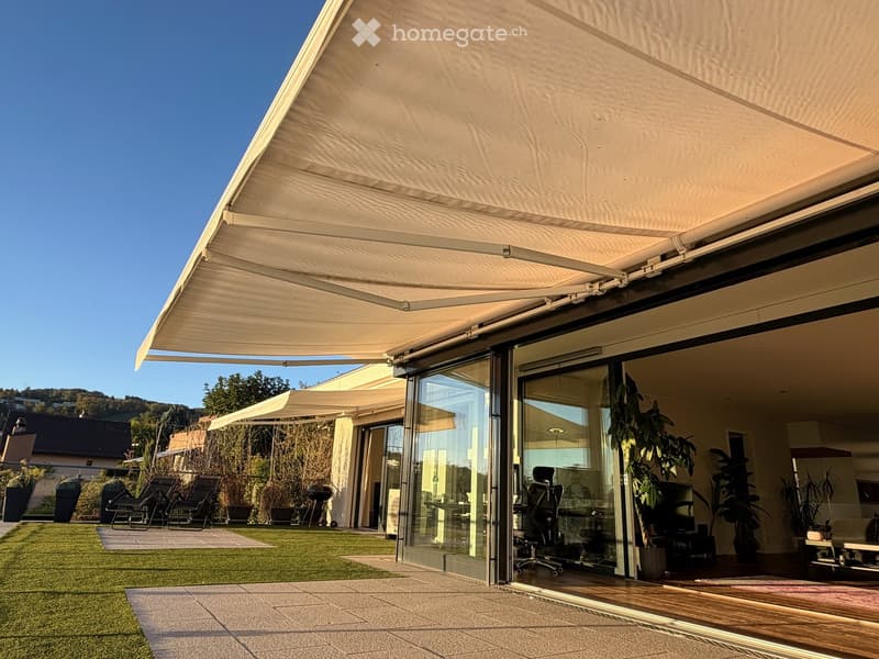 A modern outdoor patio with a large retractable awning, glass sliding doors leading to a well-maintained lawn, and comfortable seating area with chairs and a table.