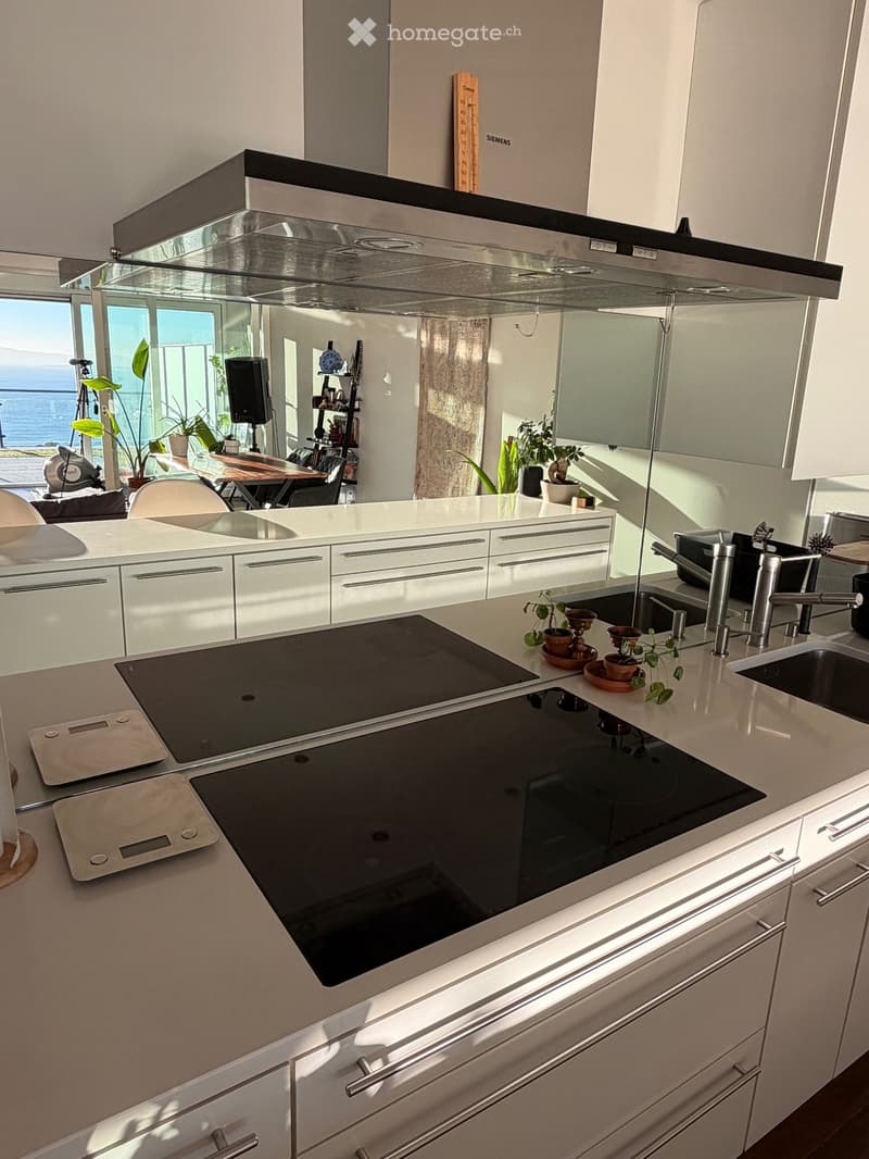 Modern kitchen with white cabinets, induction cooktop, sink, and a view of the ocean through large windows.