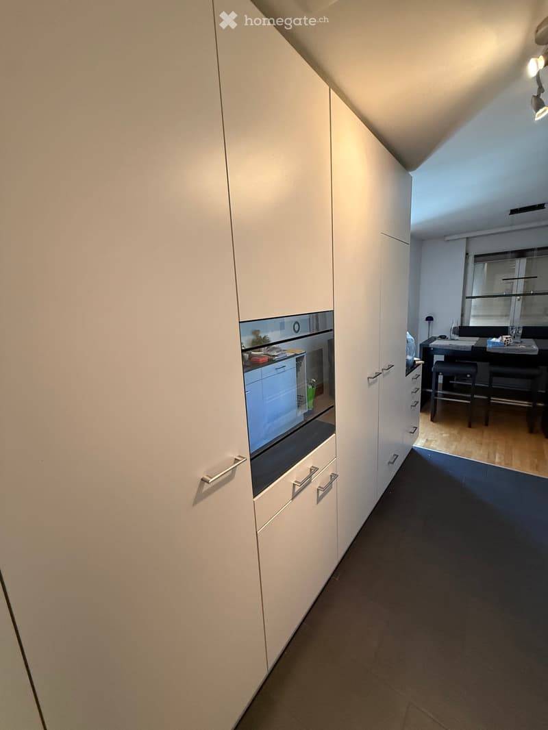 A narrow hallway with white cabinetry on one side and a glimpse of a kitchen area with an oven and countertop on the other side. The floor is dark, and the ceiling has recessed lighting.
