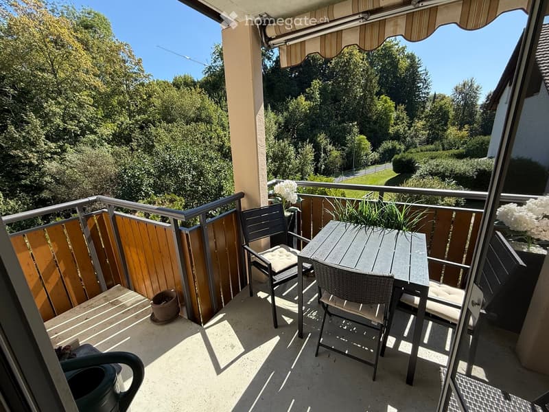 Balcony with a table, two chairs, and potted plants, overlooking a green, tree-filled area.