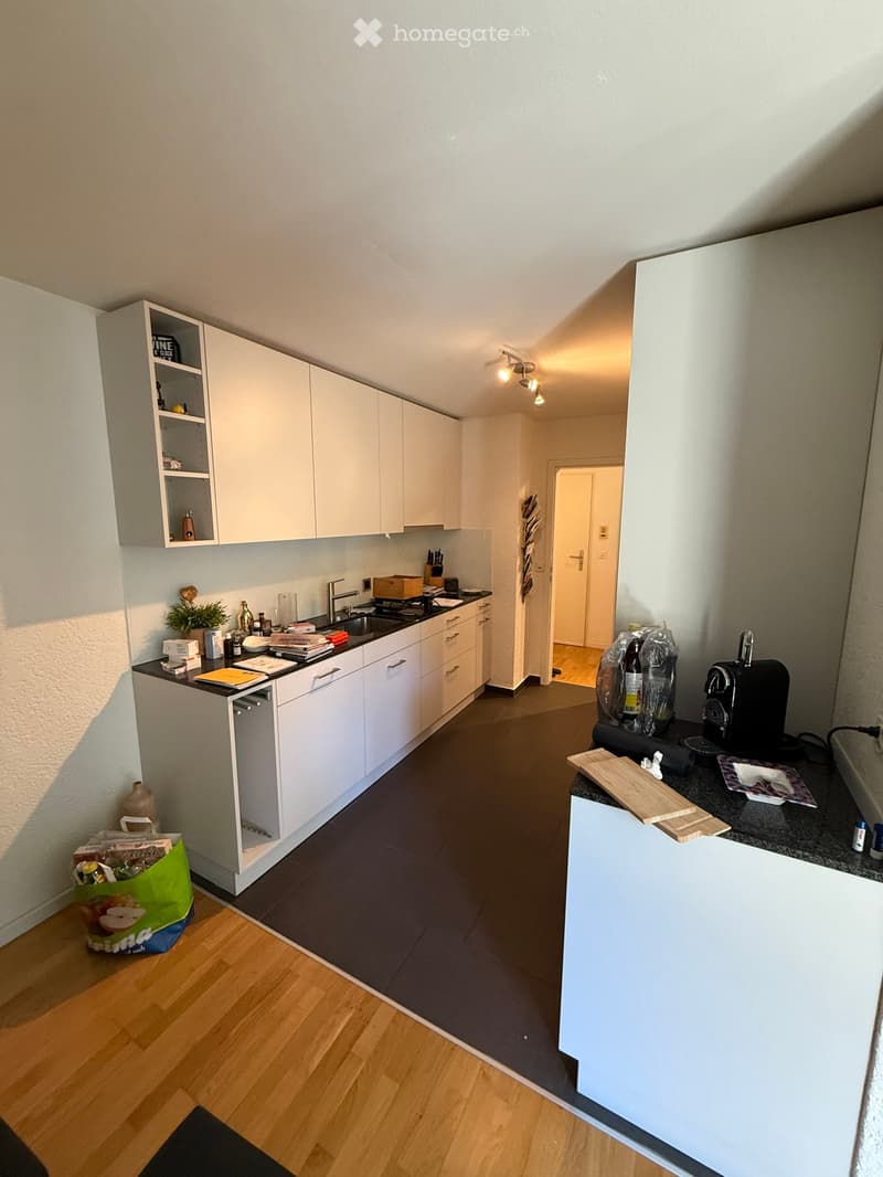 Modern kitchen with white cabinets, dark countertops, and a small island. Visible items include a coffee machine, cutting board, and a small plant on the counter.