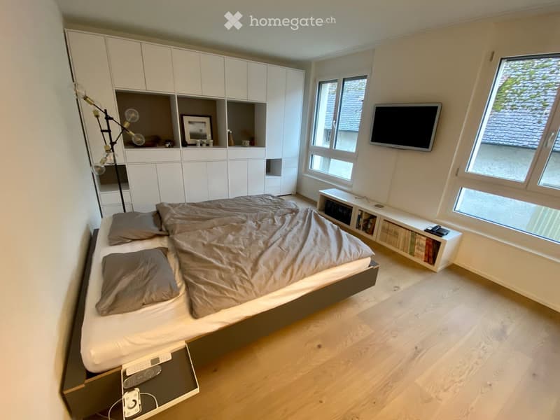 a double bed, two grey pillows, wooden floors, white walls, a cabinet, a television, a lamp