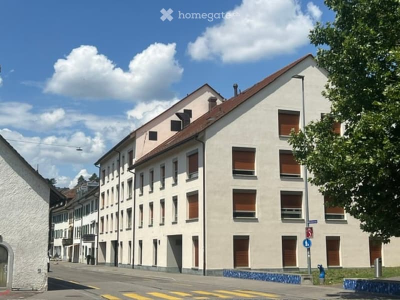 Multi-story residential building with light-colored facade, multiple windows with shutters, and a red-tiled roof.