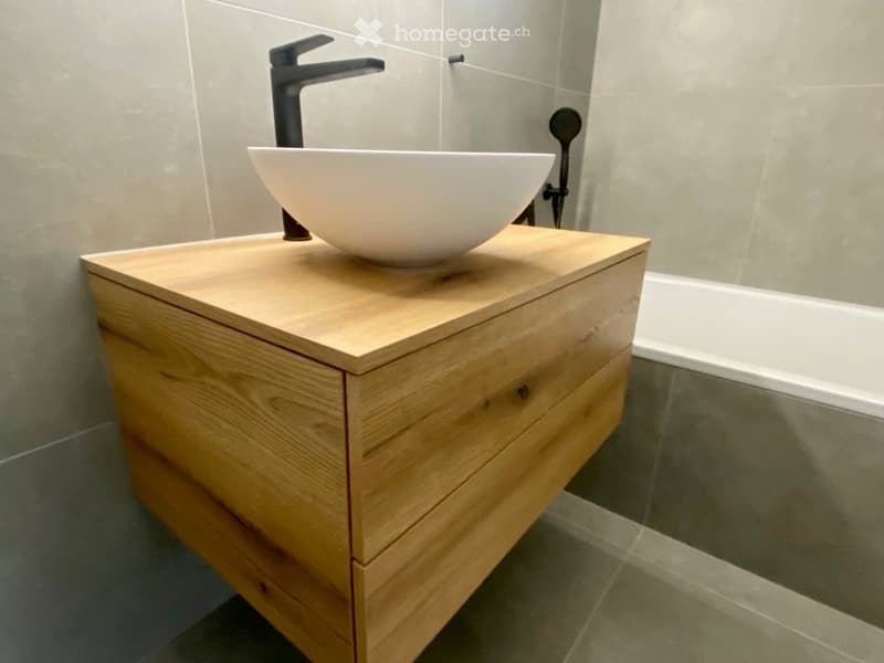 white vanity with wooden drawers, a white ceramic bowl, black faucet, a white bathtub, and tiled walls