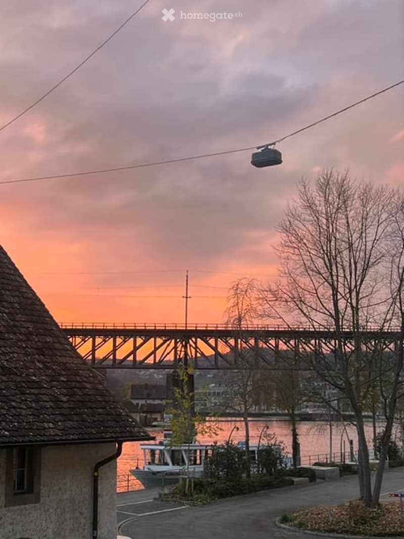 A scenic riverside view at sunset, featuring a cable car system, a bridge, and a boat on the water.