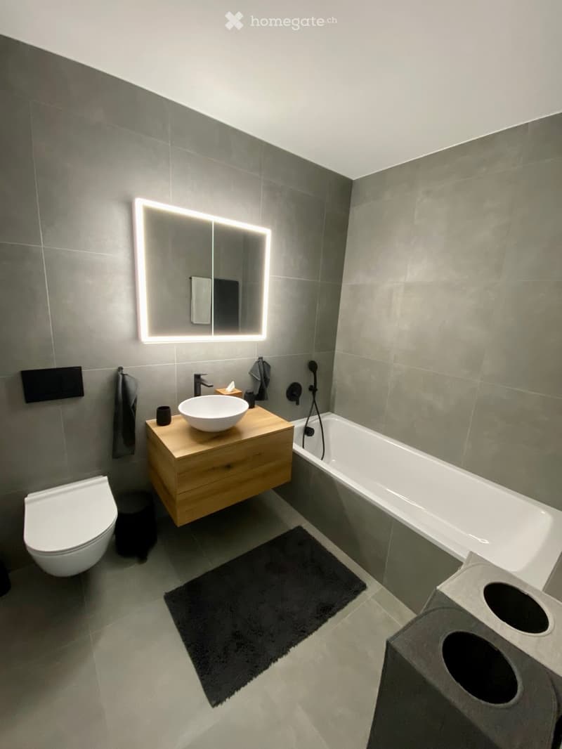 modern bathroom, white bath, silver sink, wooden vanity, grey tiles, lighted mirror, toilet, black rug, modern fixtures