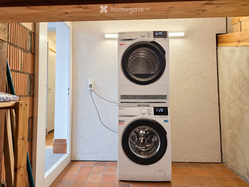 A small bathroom with a white washing machine and dryer stacked vertically, beige textured walls, and terracotta tiled floor.