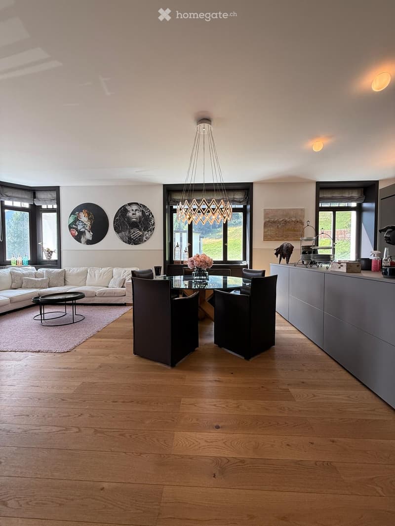 Open-plan living and dining area with light wooden flooring, white sofa, glass dining table, modern chandelier, and large windows.