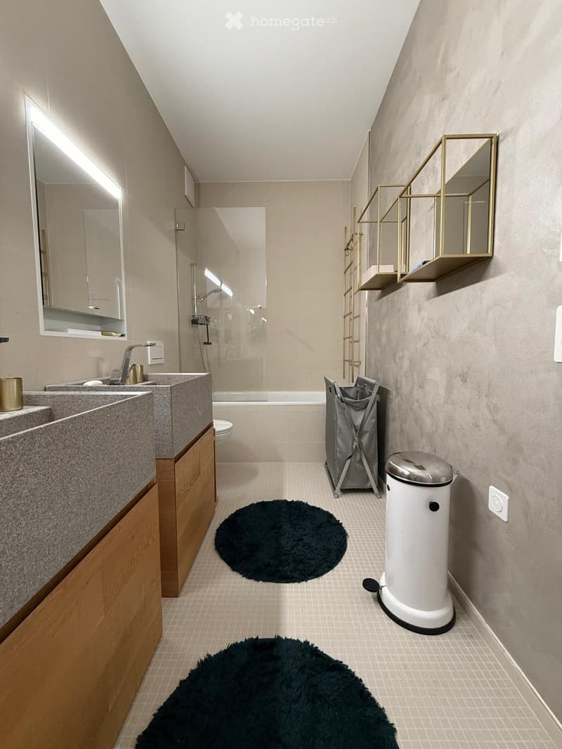 Modern bathroom with double sink, large mirror, bathtub, toilet, and built-in wooden cabinetry. Features black round rugs and wall-mounted shelves with gold accents.