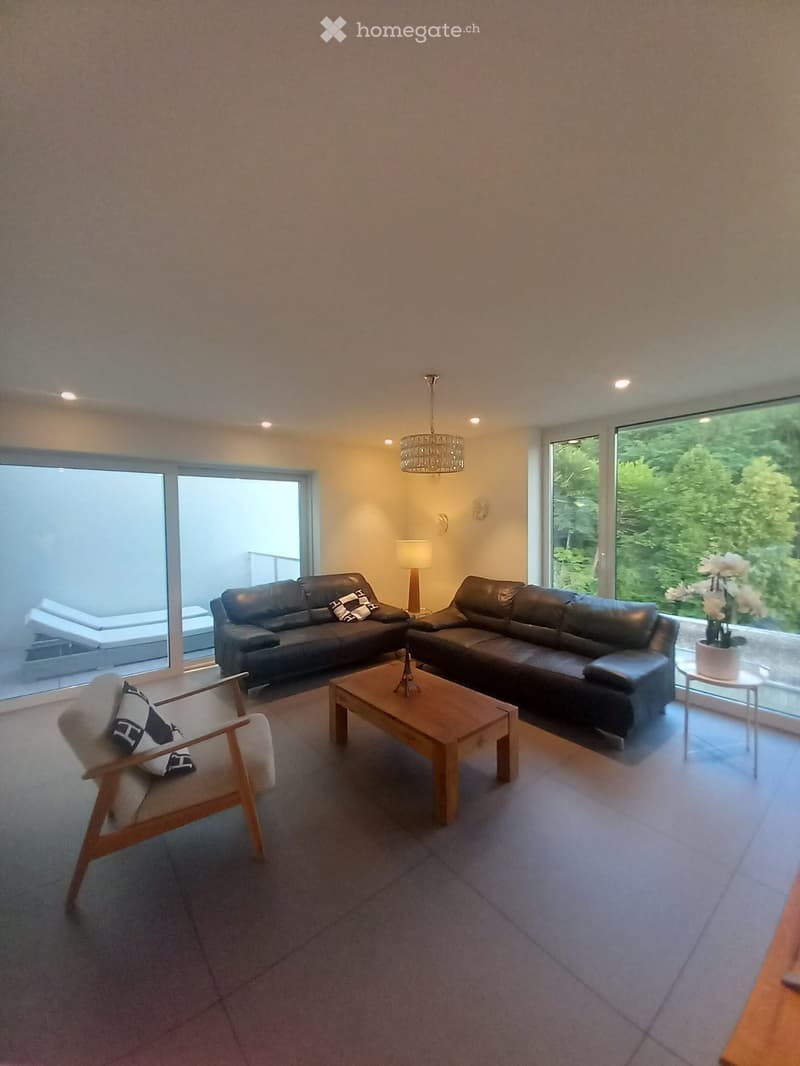 Modern living room with two black leather sofas, a wooden coffee table, a wooden armchair, a table lamp, a chandelier, and a flower pot on a side table near the large windows.