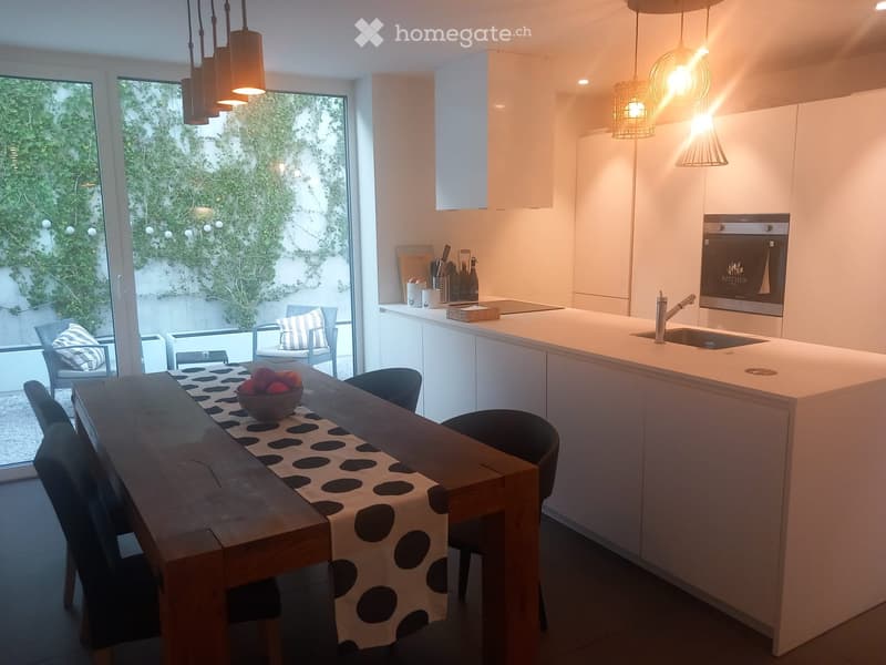 Modern kitchen with white cabinets, island, and dining area. Features a wooden dining table with chairs, a bowl of fruit, and a view of a green outdoor space.