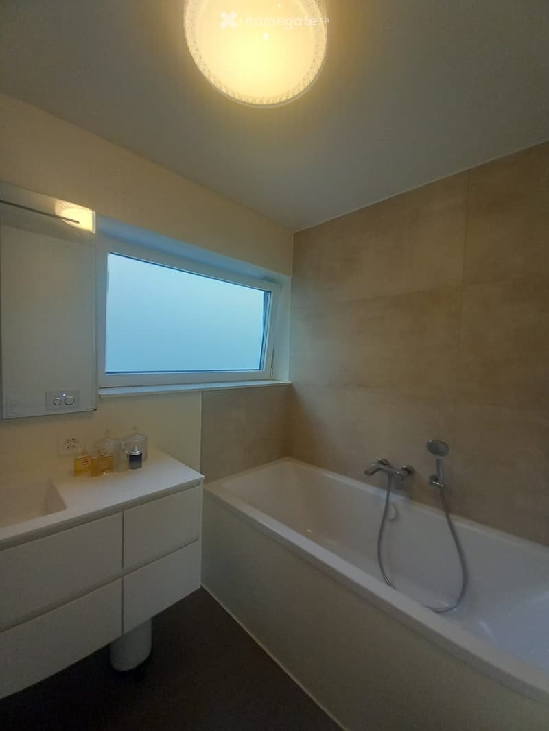 Modern bathroom with a bathtub, sink, and showerhead, featuring a large window and sleek lighting fixtures.