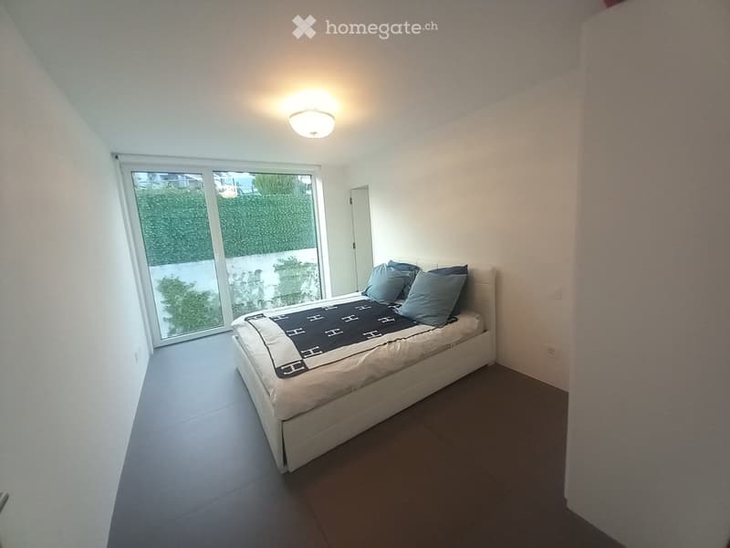 Modern bedroom with a white bed, blue pillows, and a black and white blanket, large window with a view of greenery, and a ceiling light fixture.