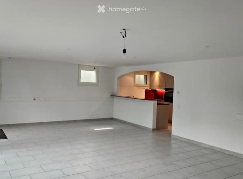 Empty room with white walls, tiled floor, and a small window. A doorway leads to a kitchen area with cabinets and appliances.