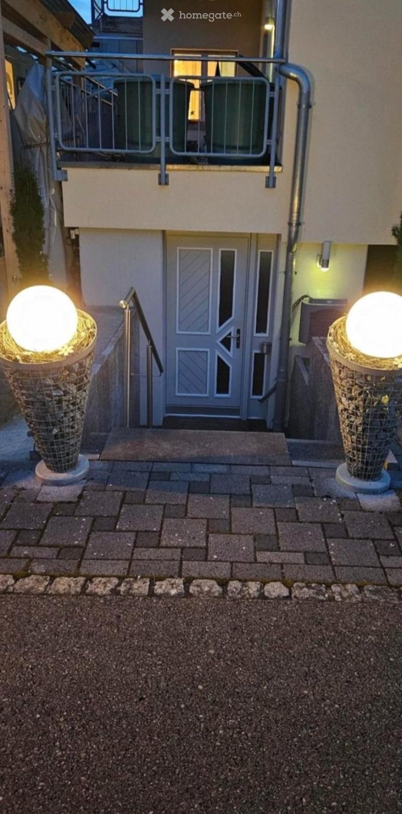 Entrance to a building with a raised ground floor, featuring a staircase leading up to a door, flanked by two spherical lamps in decorative holders, and a metal balcony above.
