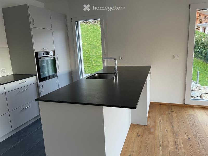 Modern kitchen with a black countertop, white cabinets, a sink with a modern faucet, and an integrated oven. The space features wooden flooring and large windows offering a view of the garden.
