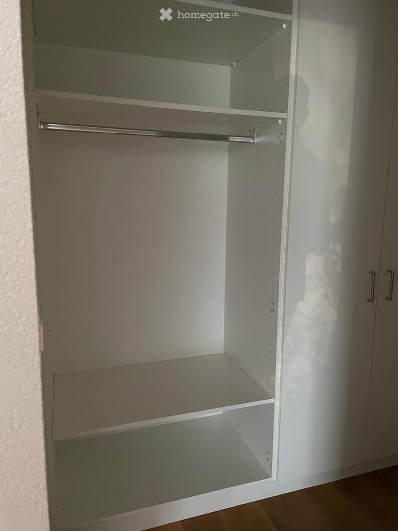 Empty closet with multiple shelves and a hanging rod, white walls and wooden floor visible.