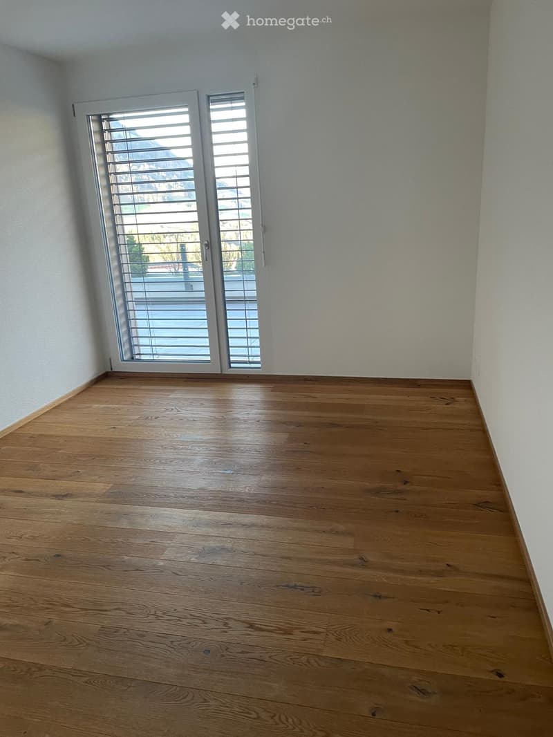 Empty room with wooden flooring and large windows with blinds, allowing natural light to enter.