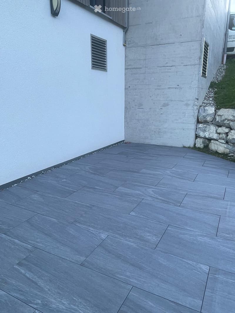 Grey tiled patio area adjacent to a white building with a stone retaining wall and small grassy area.