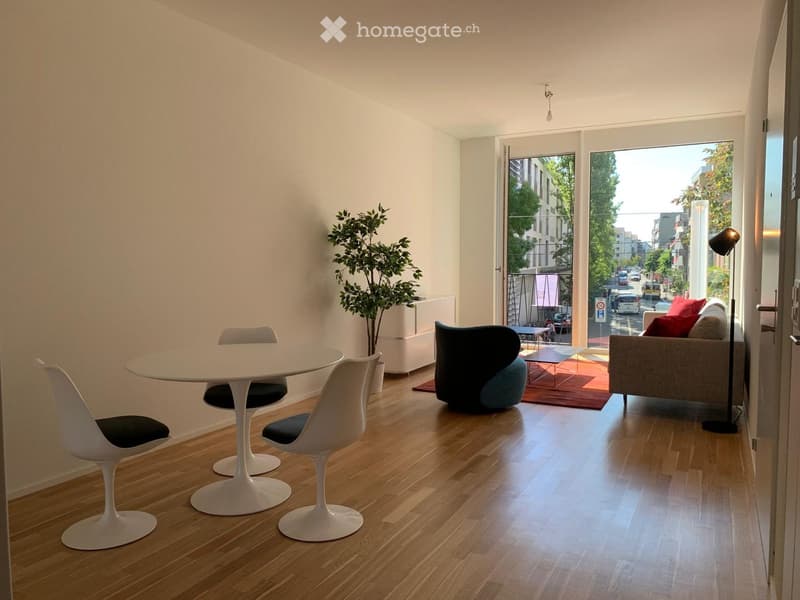 Modern living room with wooden flooring, white dining table with chairs, grey sofa with red cushions, floor lamp, and potted plant near the window.
