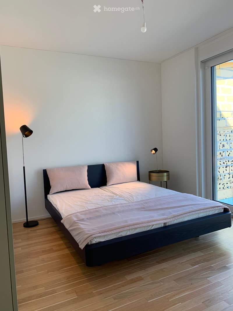 Modern bedroom with a double bed, nightstand, and floor lamp. The room features wooden flooring and large windows with sheer curtains.